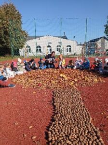 Na zdjęciu dzieci siedzące na boisku szkolnym. Przed dziećmi widoczne drzewo z usypanych darów jesieni.
