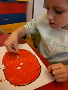 Na zdjęciu chłopiec siedzący przy stoliku i dekorujący swoją dynię pestkami z dyni.