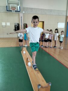 Na zdjęciu chłopiec na sali gimnastycznej chodzący po ławeczce i ćwiczący utrzymywanie równowagi.
