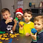 Czterech chłopców ( chopiec w bluzie z białym napisem, chłopiec w czapce Mikołaja, chłopiec w limonkowej bluzie oraz chopiec w bluzie z zielonymi wstawkami) w swoich rączkach pokazują ukończone ciasteczko piernikowe.