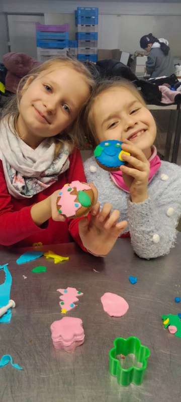 Dwie dziewczynki, które się uśmiechają i podnoszą do zdjęcia swoje kolorowe gotowe ciasteczko piernikowe. Jedna dziewczynka ma czerwony sweter, a druga szary z białymi kuleczkami.