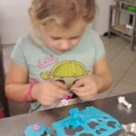 Pochylona dziewczynka wykrawa różne wzory w swojej niebieskiej masie cukrowej.