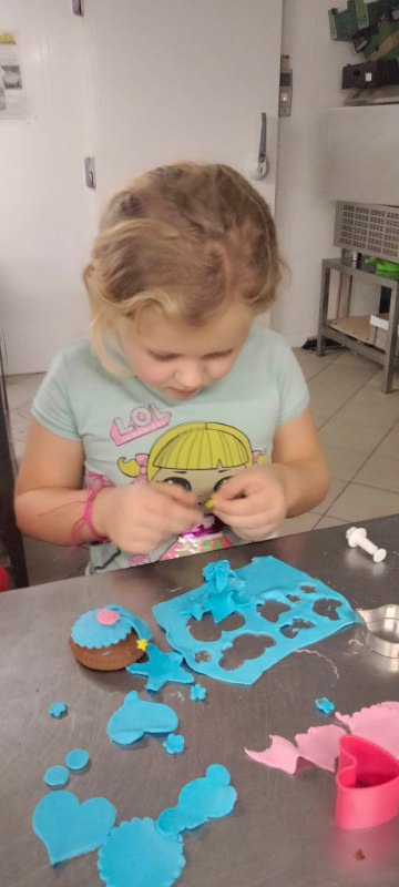 Pochylona dziewczynka wykrawa różne wzory w swojej niebieskiej masie cukrowej.