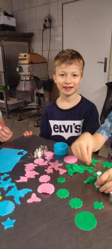 Widać chłopca w granatowej bluzce z dużym napisem na białym tle -eLvis. Przed nim na stole widać masę cukrową w kolorze różowym, po prawej stronie masę zielona i po lewej niebieską.