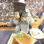 Chłopiec ubrany w strój pszczelarza trzyma w ręku podkurzacz i słoik miodu.