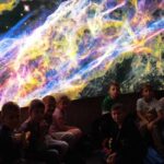 Grupa chłopców siedzaca na tle podświetlonego plakatu galaktyki.