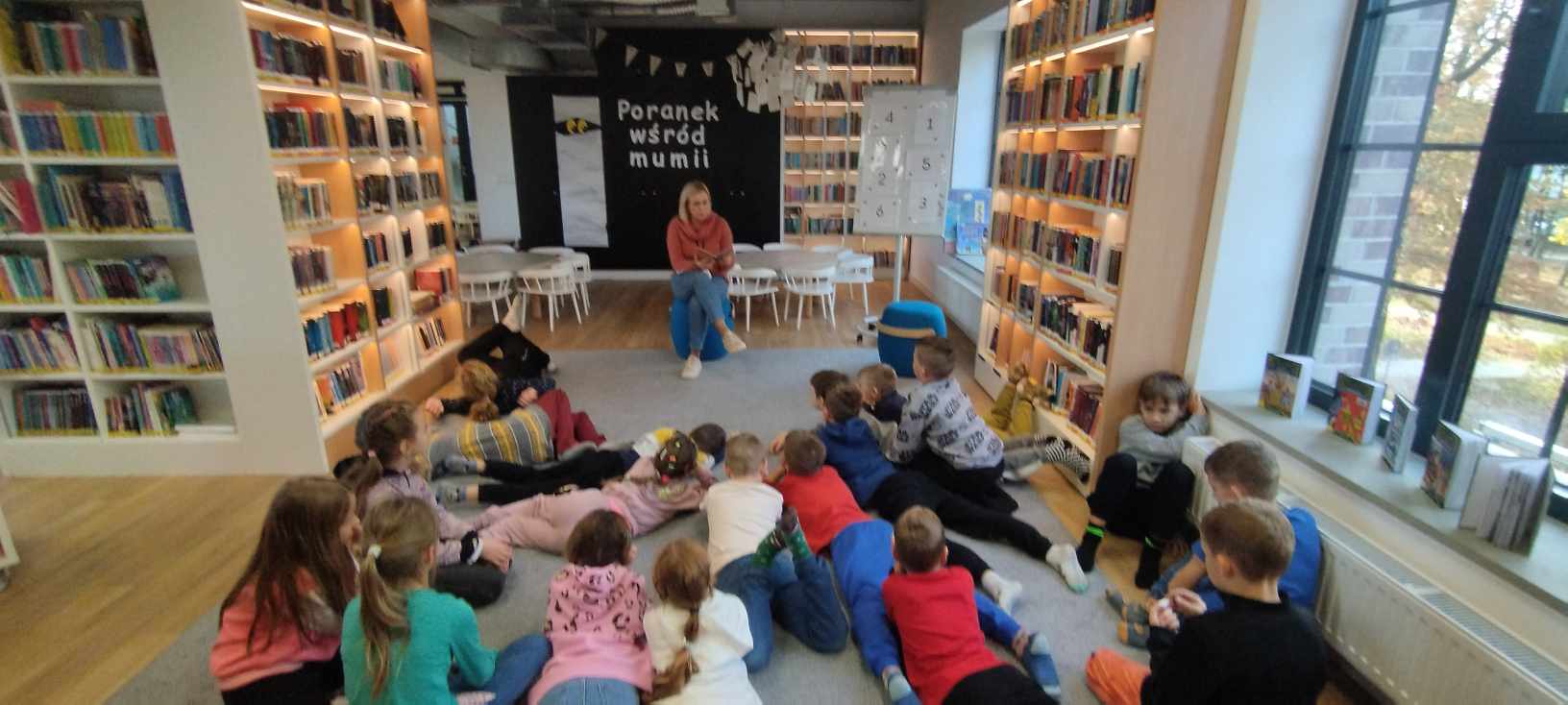 Pani Bibliotekarka czyta fragment książki. Na dywanie przed siedzą i leżą dzieci.