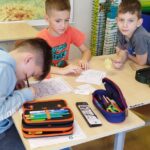 Trzech chłopców siedzi przy stoliku i rozwiązuje test i zagadki. Chłopiec w szarej bluzie pisze. W tle widać mapę Polski.