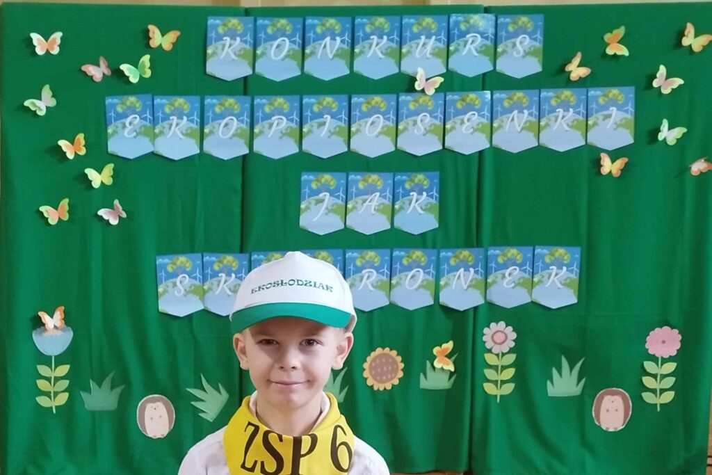 Zdjęcie twarzy chłopca na tle zielonej dekoracji, z niebieskimi literami - konkurs ekopiosenki jak skowronek. Chłopiec stoi w białej czapce z zielonym daszkiem, na szyi ma zawiązaną żółtą chustkę - ZSP6 w Żorach.